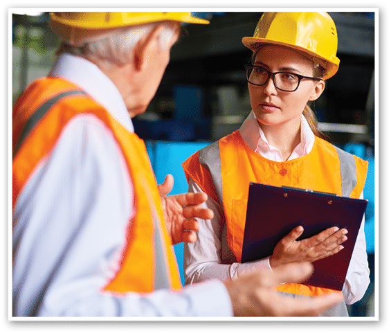 Pretty young factory worker and her senior colleague discussing results of accomplished work while standing at production department, both of them wearing hardhats and reflective jackets